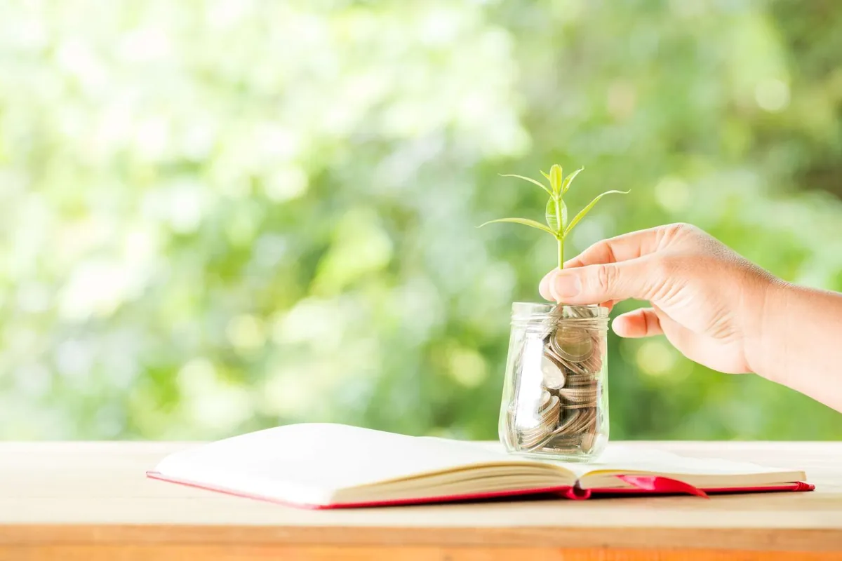 A hand placing a small green plant into a jar of coins on an open notebook