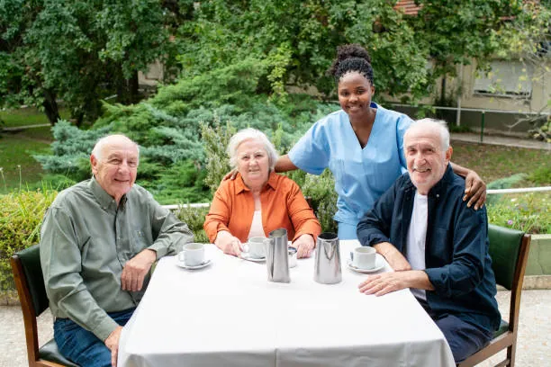 Three older adults sitting at a garden table with tea, with a smiling care worker standing behind them